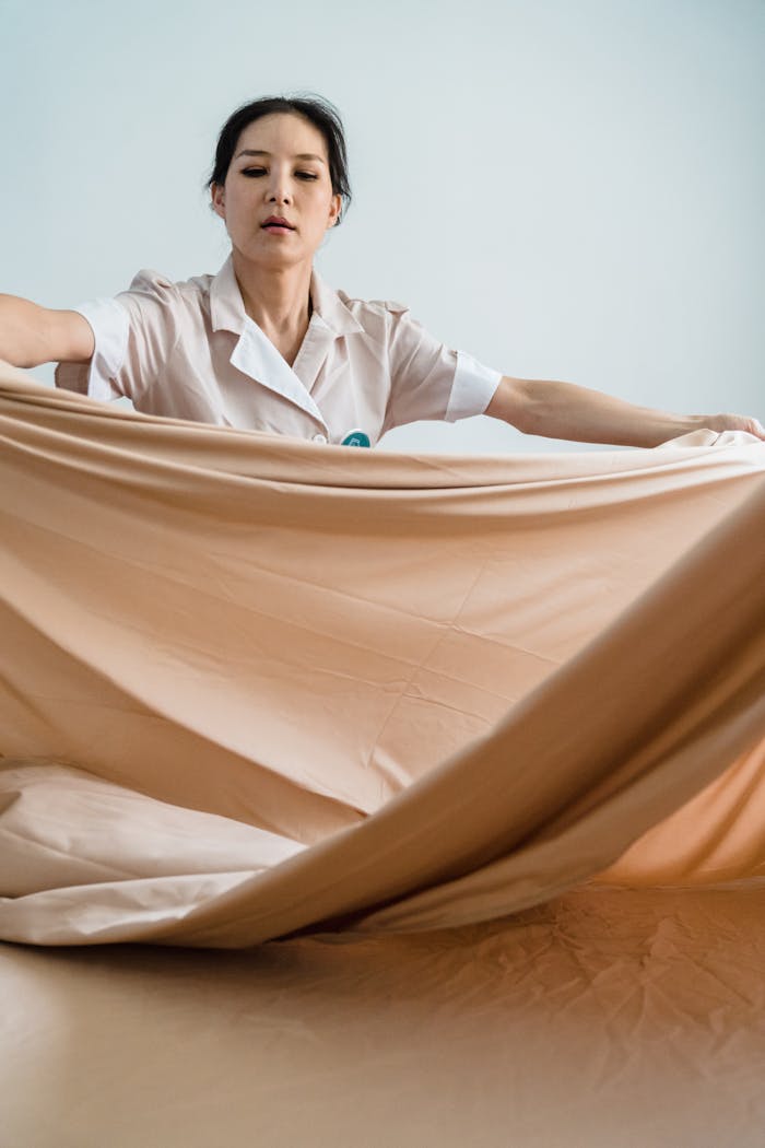 A woman in uniform making a bed with beige sheets, showcasing housekeeping services.